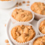 Five peanut butter chip muffins in white paper liners are arranged on a white plate, with a bowl of peanut butter and scattered chips in the background.