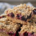 Three crumbly lemon blueberry bars are stacked on a plate, with a blurred checkered cloth in the background.