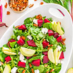 A white dish with a salad containing arugula, sliced green apples, chunks of beetroot, goat cheese, and pecans. Loose pecans and beet greens are scattered around.