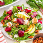 A plate of arugula salad with apple slices, roasted beets, pecans, and crumbled goat cheese on a white surface.