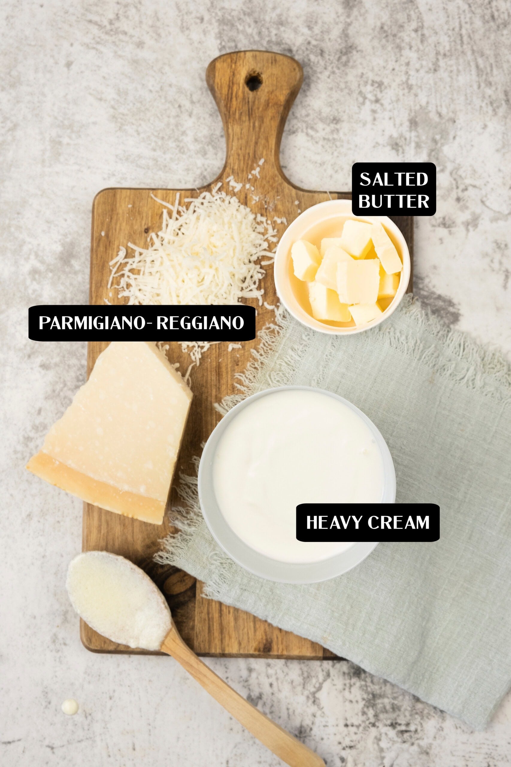 A wooden board displays grated Parmigiano-Reggiano, a bowl of salted butter cubes, a wedge of cheese, a bowl of heavy cream, and a spoon of grated cheese—perfect ingredients for an authentic alfredo sauce recipe.