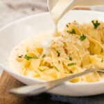 A white sauce is being poured from a jug onto a bowl of fettuccine pasta, garnished with grated cheese and chopped parsley.
