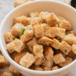 A white bowl filled with seasoned croutons sits on a surface scattered with more croutons, a small dish of chopped herbs nearby—perfect as one of the game day foods that you can make in the air fryer.