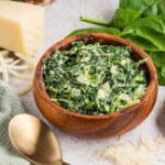 A wooden bowl filled with creamy spinach sits on a table surrounded by fresh spinach leaves, garlic cloves, shredded cheese, and a piece of cheese.