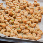 A baking tray lined with parchment paper holds evenly spread, golden-brown homemade croutons. Metal tongs and fresh herbs are visible nearby.