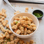 A white bowl filled with seasoned croutons, surrounded by more croutons on parchment paper, with metal tongs and a small dish of chopped herbs nearby.