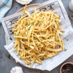 A plate of seasoned shoestring French fries garnished with chopped herbs, served on newspaper with a striped towel, garlic, pepper, and a salt shaker nearby.
