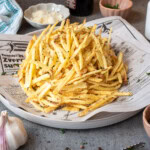 A pile of thin, seasoned French fries sits on newspaper atop a plate, surrounded by bowls of peppercorns, grated cheese, chopped herbs, a garlic bulb, and a salt shaker.