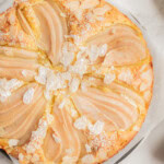 A round pear almond cake topped with sliced pears, almond flakes, and powdered sugar, displayed on parchment paper.