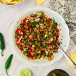 A bowl of chopped salsa with tomatoes, onions, and herbs sits on a marble surface, surrounded by pickles, lime halves, jalapeños, tortilla chips, and a patterned cloth.