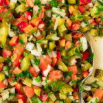 A close-up of chopped salad with tomatoes, onions, pickles, and herbs in a white bowl, with a metal spoon partially visible.