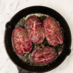 Four pieces of raw beef are cooking in a cast iron skillet on a white marble countertop.
