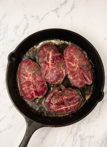 Four pieces of raw beef are cooking in a cast iron skillet on a white marble countertop.
