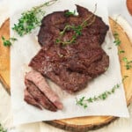 Two pieces of cooked steak with herbs on parchment paper, placed on a round wooden board, with a small bowl of pink salt nearby.