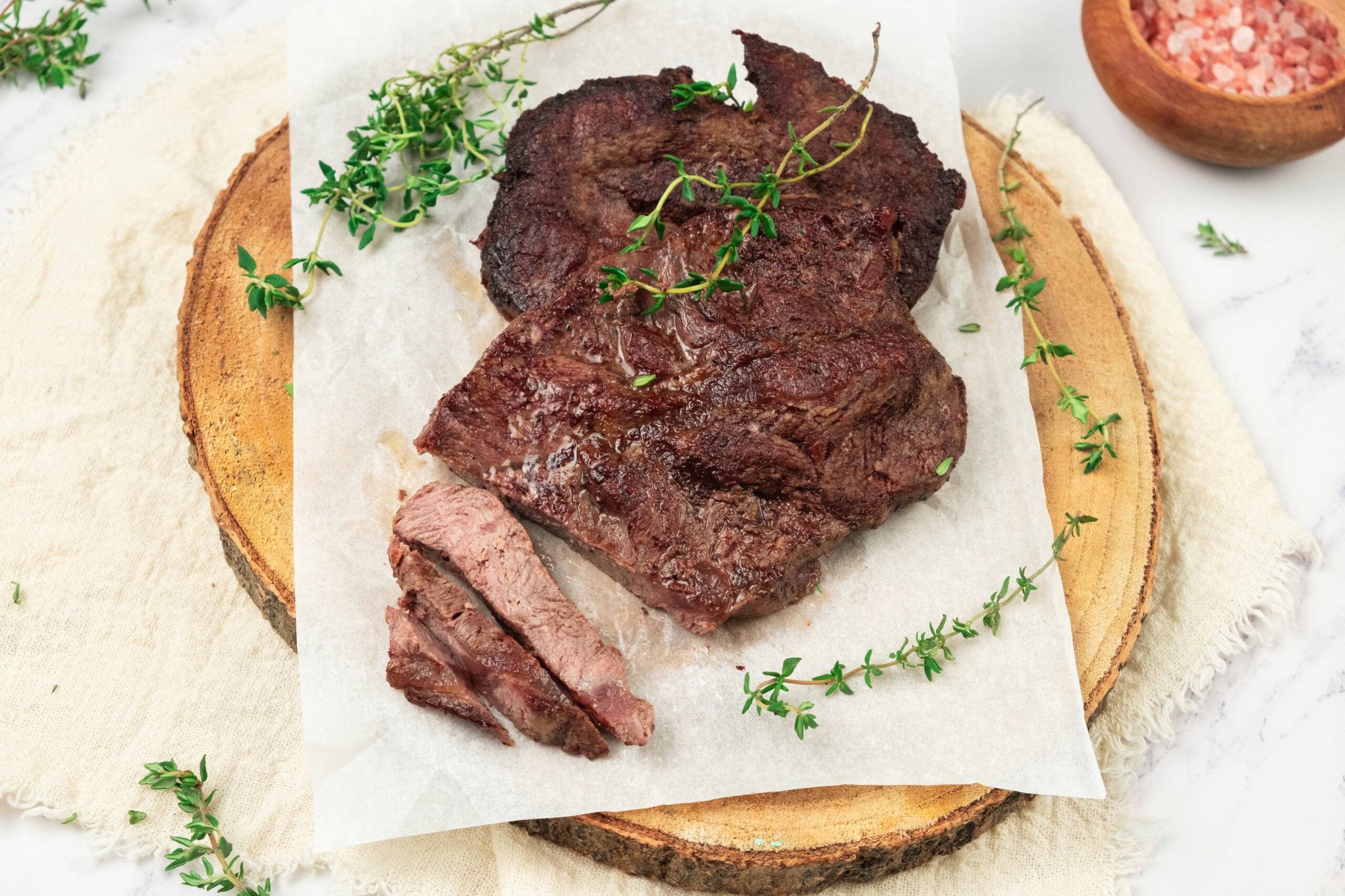 Two pieces of cooked steak with herbs on parchment paper, placed on a round wooden board, with a small bowl of pink salt nearby.