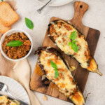 Stuffed eggplants topped with melted cheese and fresh basil on a wooden board, served with extra filling and toast on the side.