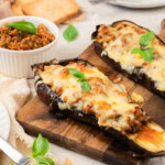 Two baked eggplant halves stuffed with meat and melted cheese, garnished with basil, served on a wooden board with toast and a small bowl of spread nearby.