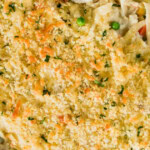 A baked chicken casserole topped with golden breadcrumbs and herbs, featuring visible pieces of vegetables and noodles in a white dish.