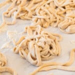 Uncooked, hand-cut noodles are spread out on a floured surface, ready to be cooked.