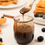 A spoon drizzling syrup into a small glass jar, surrounded by waffles, blueberries, and cinnamon sticks on a white surface.