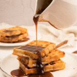 A hand pours cinnamon roll syrup from a white pitcher onto a stack of waffles on a white plate, with more waffles on a plate in the background.