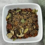 A white square bowl filled with a mixture of whole and crushed spices, including bay leaves, dried chili, peppercorns, and seeds, on a gray surface next to a green cloth.