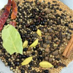 A metal bowl containing whole spices including dried red chilies, bay leaf, cardamom pods, mustard seeds, cloves, black peppercorns, allspice, and a cinnamon stick.