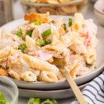 A plate of creamy pasta salad with penne, imitation crab meat, and chopped green onions, served with a fork.