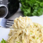 A fork holds a bite of creamy shredded chicken pasta, with a plate of the same dish and fresh parsley in the background.