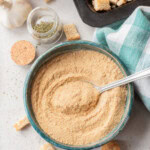 A bowl of breadcrumbs with a spoon, next to croutons in a baking tray, a garlic bulb, a jar of dried herbs, and a green checkered cloth on a white surface.