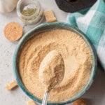 A spoonful of breadcrumbs held over a bowl, with croutons, a jar of dried herbs, a garlic bulb, and a checkered cloth nearby on a light surface.