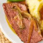 Two slices of pot roast topped with onions and gravy, served with mashed potatoes and green beans on a white plate.