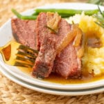 Sliced roast beef with onions and gravy, served with mashed potatoes and green beans on a white plate with a gold fork.