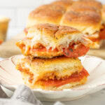 Two stacked slices of baked sliders filled with pepperoni and melted cheese on a white plate, with more sliders in the background.