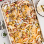 Sheet pan with baked chicken breasts, baby potatoes, onions, and apple slices on foil, surrounded by side dishes and utensils on a white table.