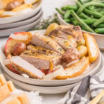 A plate of pork chops and apples is served with roasted potatoes, onions, and apple slices. Green beans, bread, and seasonings are visible in the background.