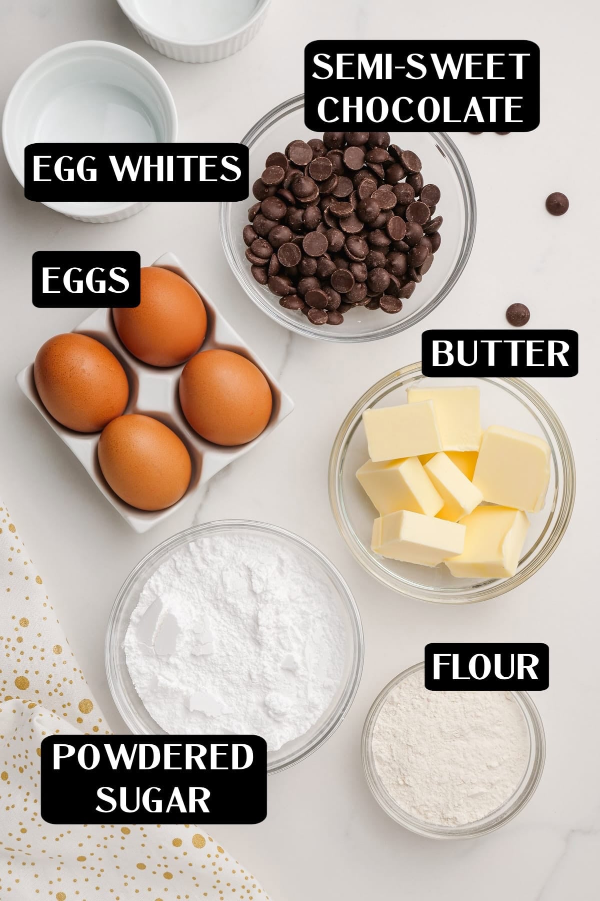 molten lava cake 1 Top-down view of baking ingredients for molten Lava cakes: egg whites, eggs, semi-sweet chocolate chips, butter cubes, powdered sugar, and flour in separate bowls on a white surface.