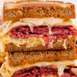 A classic Reuben sandwich stacked with corned beef, sauerkraut, melted Swiss cheese, and dressing on rye bread, served with a pickle spear.