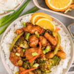 A plate of white rice topped with stir-fried chicken, broccoli, bell peppers, and green onions, garnished with orange slices. Nearby are rice, a sliced orange, and chopsticks.