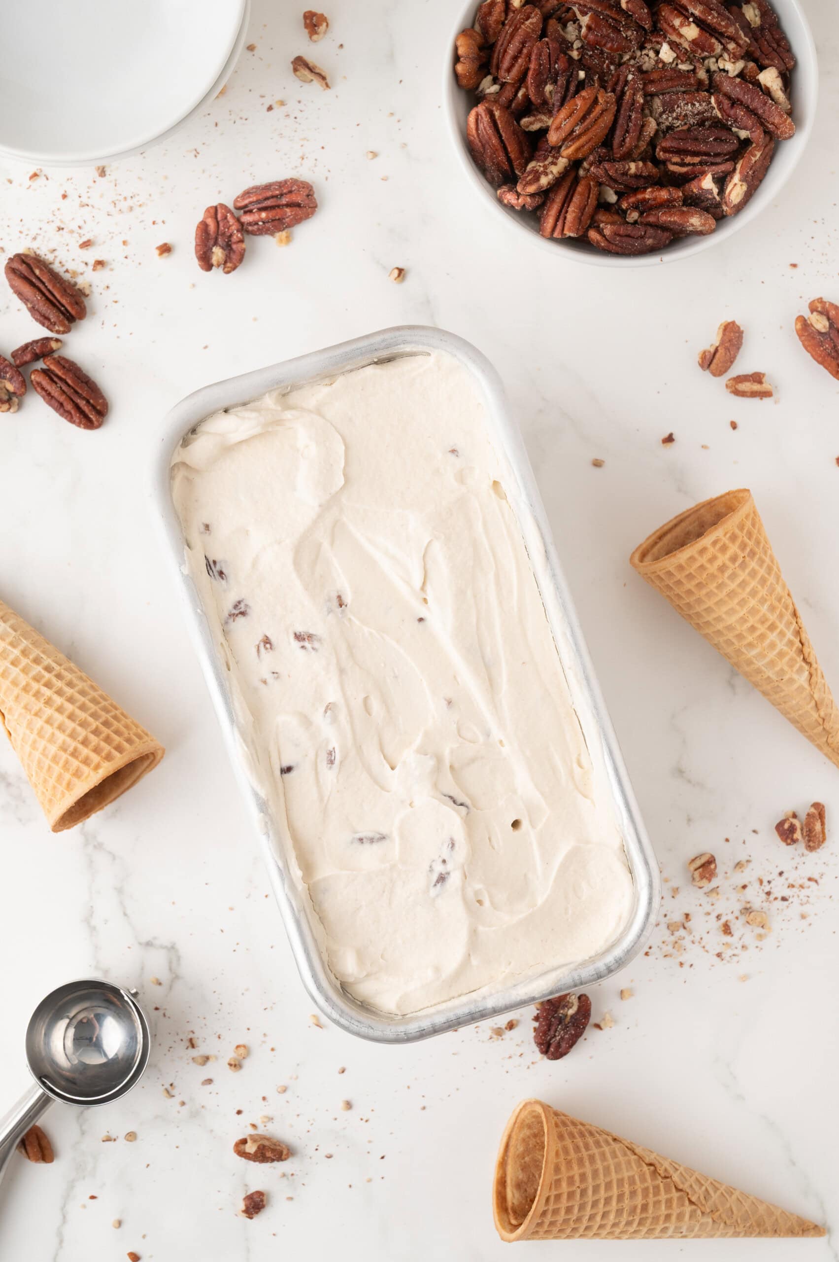Rectangular container of pecan ice cream on a white surface with pecans, empty waffle cones, and a metal ice cream scoop scattered around.