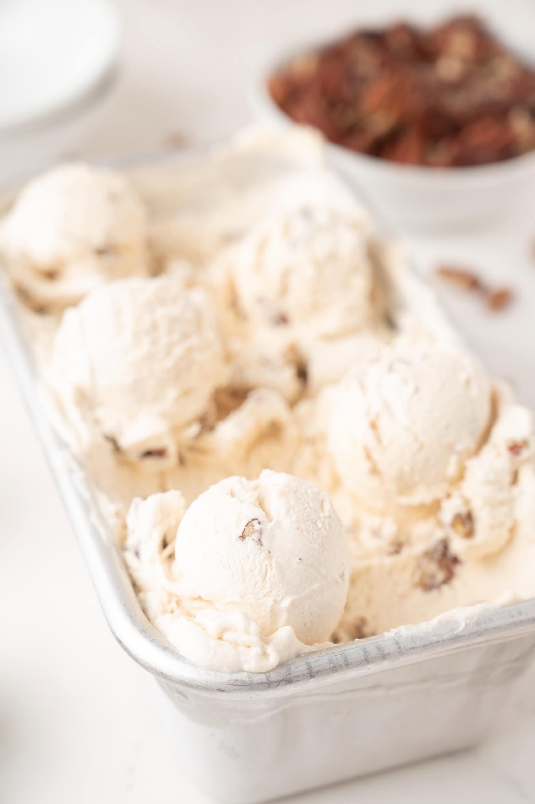 A metal container filled with several scoops of pecan ice cream, with a blurred bowl of pecans in the background.