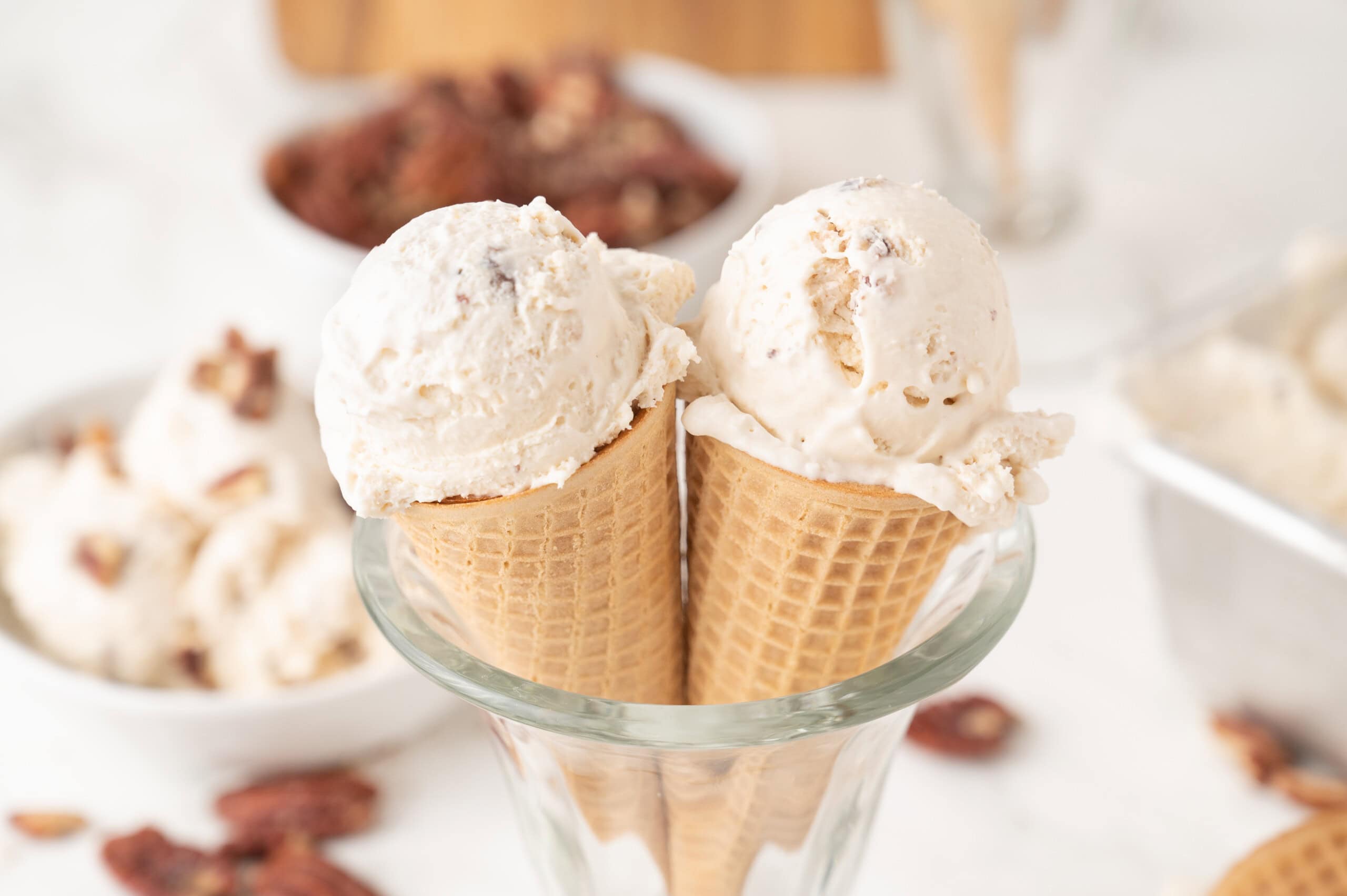 Two waffle cones each hold a scoop of pecan ice cream, placed in a glass. Bowls of pecans and more ice cream are in the background.