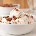 A white bowl filled with scoops of vanilla ice cream topped with pecans, with blurred bowls and cones in the background.