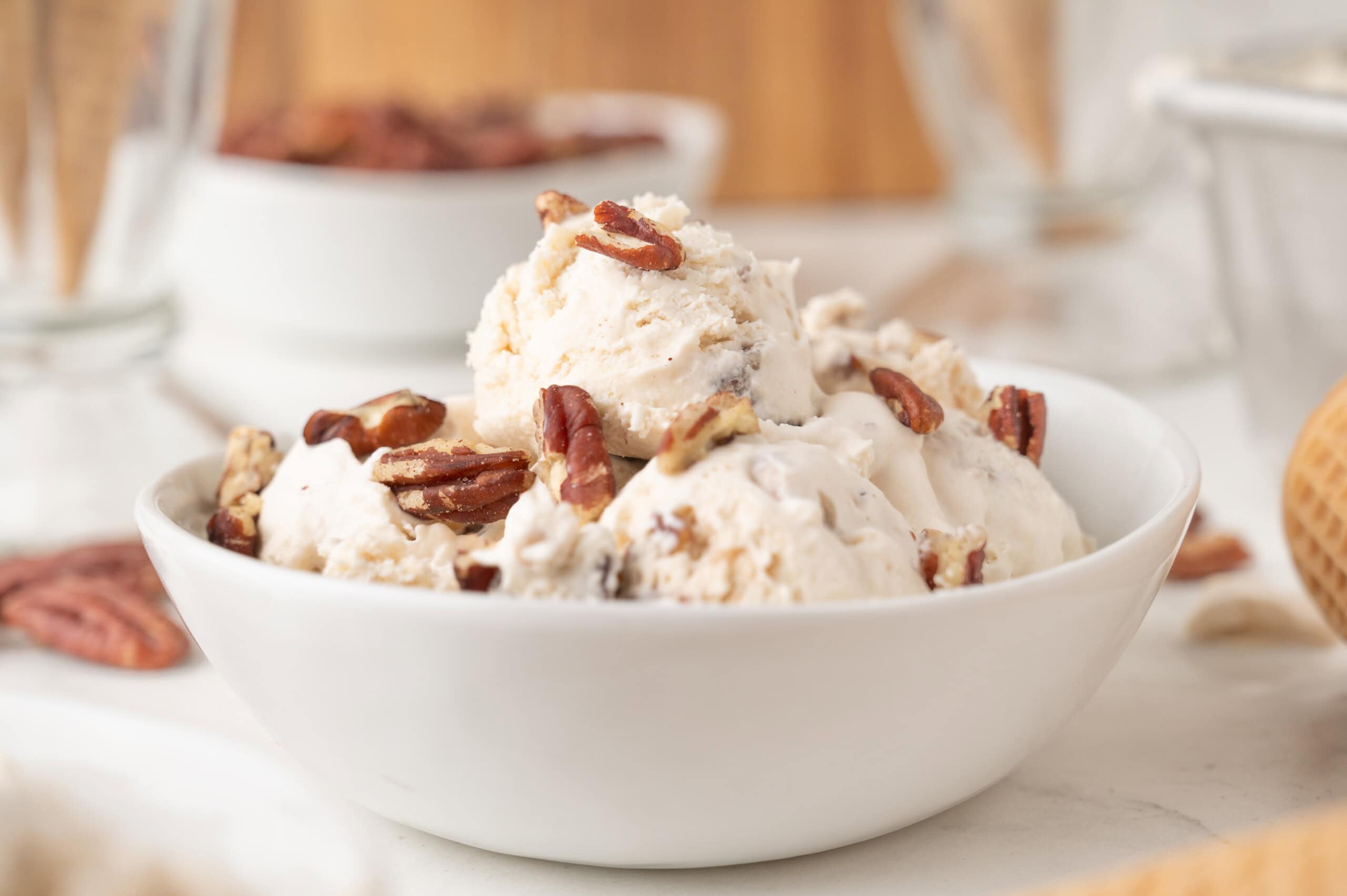 A white bowl filled with scoops of vanilla ice cream topped with pecans, with blurred bowls and cones in the background.