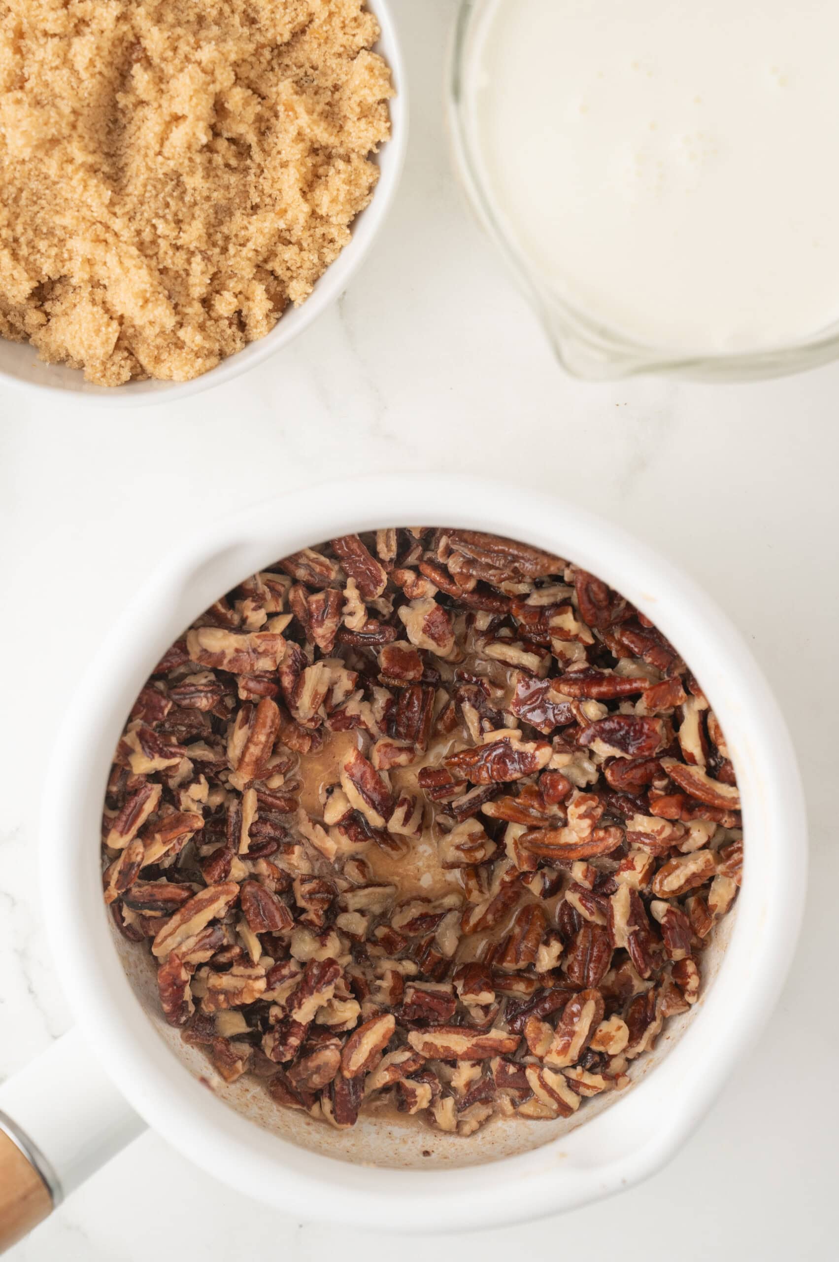 Overhead view of a saucepan with chopped pecans, a bowl of brown sugar, and a pitcher of cream on a white surface.