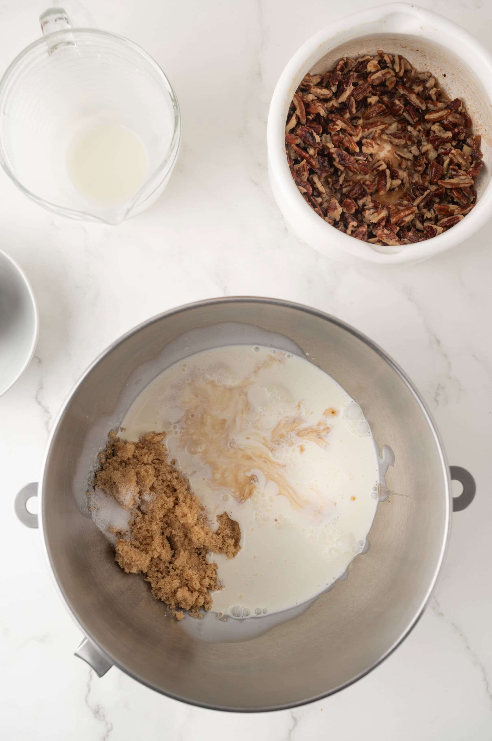 A metal mixing bowl with brown sugar, milk, and vanilla, a bowl of chopped pecans, and a glass measuring cup with milk, all on a white countertop.
