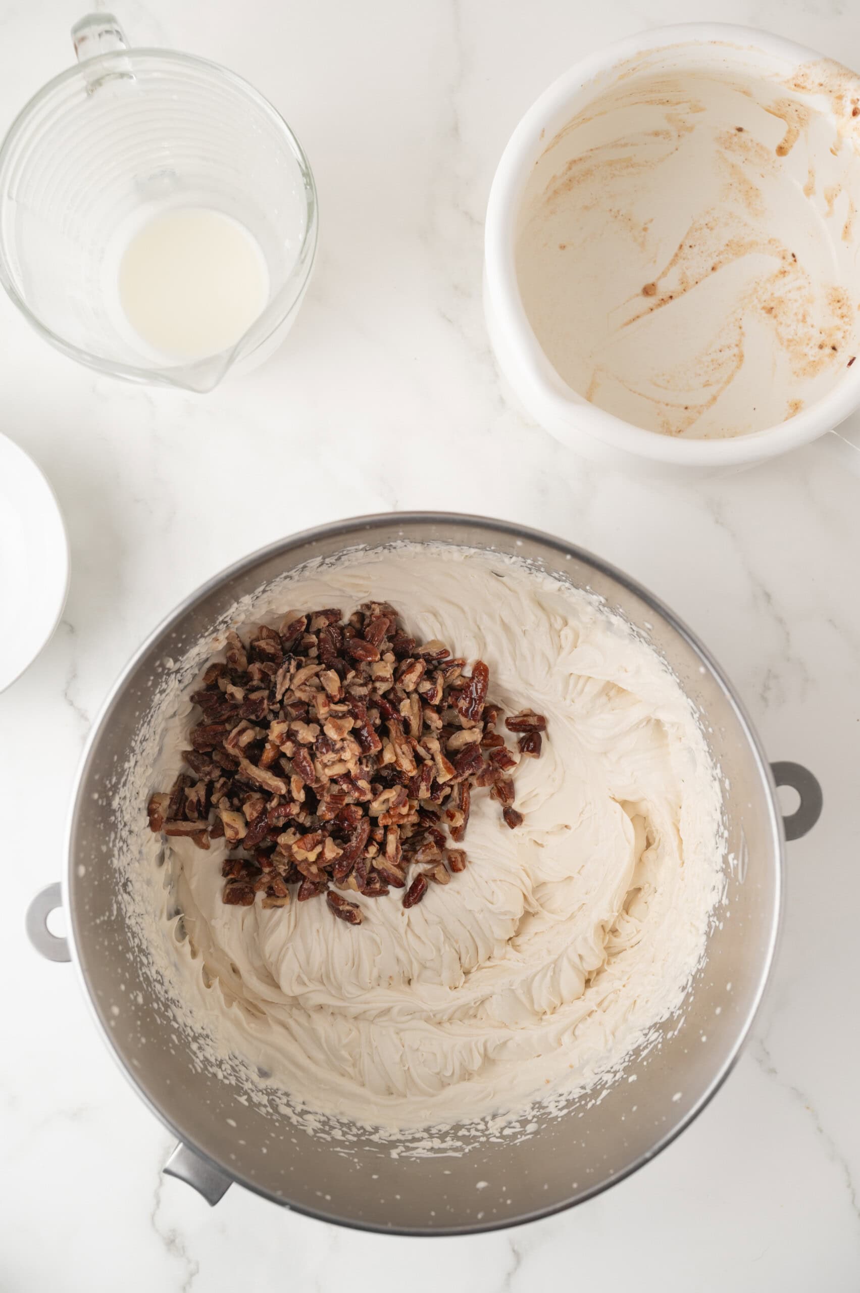 A metal mixing bowl with whipped ice cream base and chopped pecans on top, next to an empty bowl with frosting residue and a glass measuring cup with milk on a white countertop.