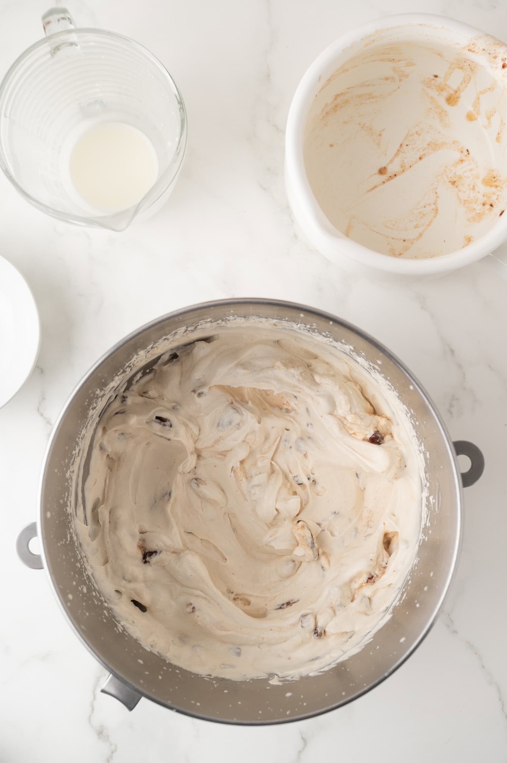 A mixing bowl filled with creamy, mixed cookie dough, with a jug of milk and an empty mixing bowl nearby on a white countertop.