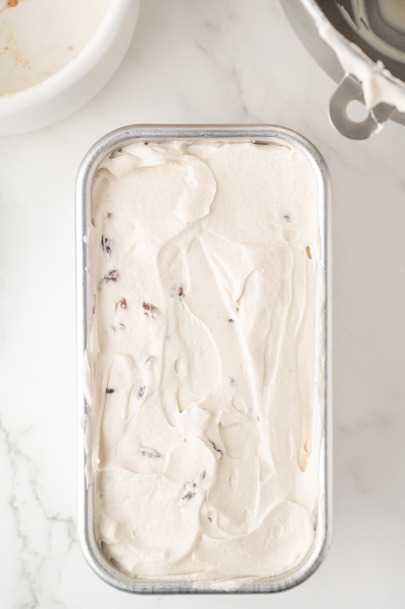 A metal container filled with creamy ice cream mixture with visible chocolate or fruit pieces on a white surface.