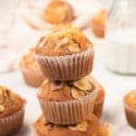 Three almond-topped muffins are stacked on top of each other, surrounded by more muffins on a white surface, with a glass bottle of milk in the background.